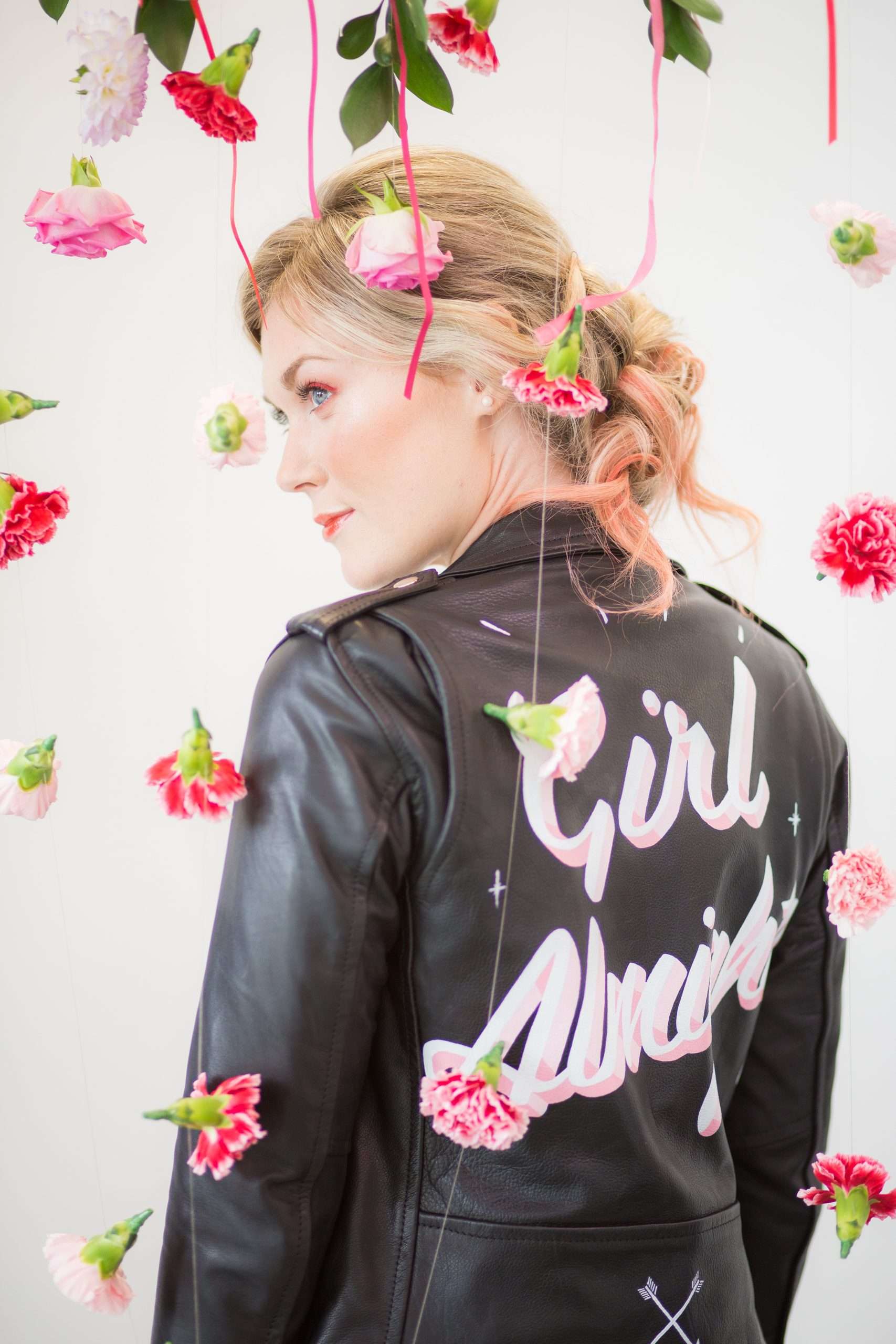 Girl in Laurie Lee independent designer leather jacket with Girl Almighty painted on back, standing behind curtain of pink flower heads