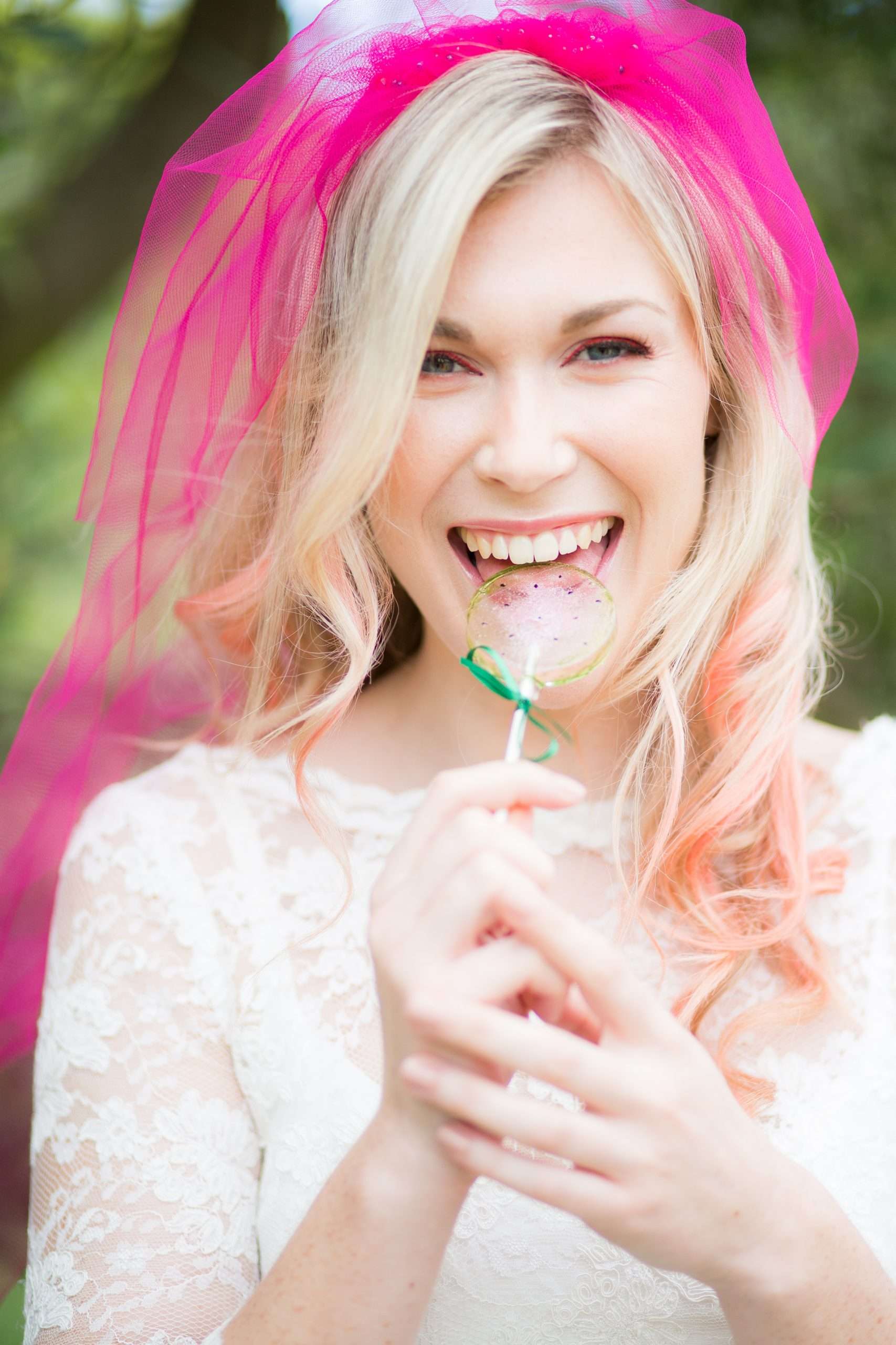 Blonde model modelling wedding dress in white lace dress and shocking pink veil smiling brightly into camera while eating a transparent pale pink and watermelon green lollipop tied with a green ribbon