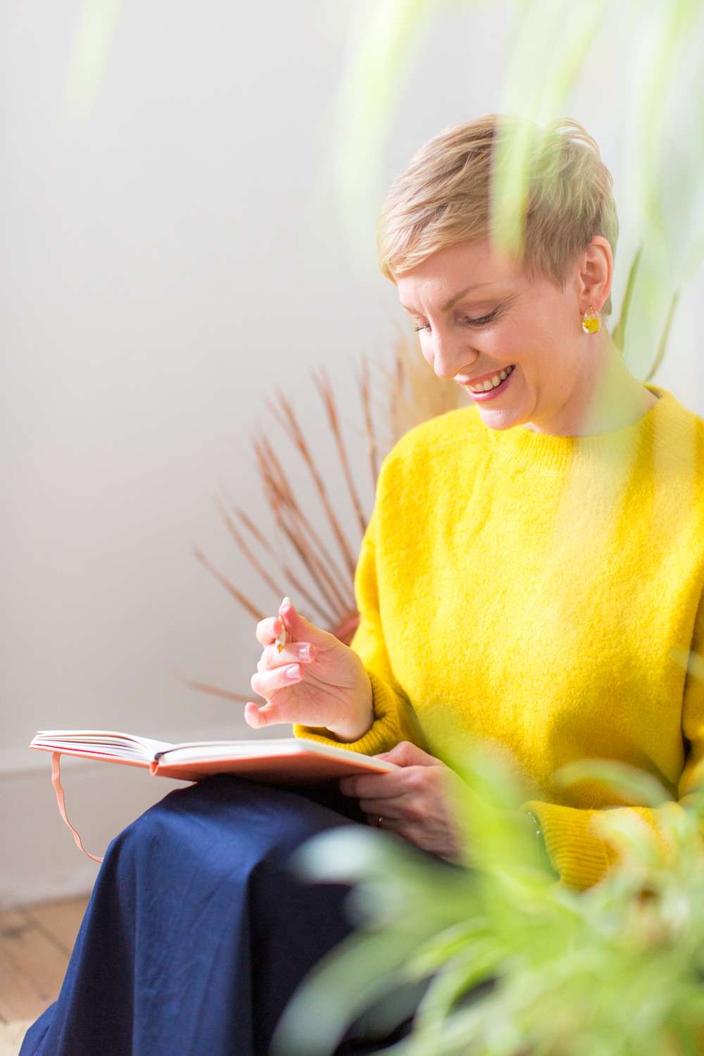 coach dressed in bright yellow jumper and navy skirt sitting down and journalling - we are peeking through foreground blurred leaves at her, there's a palm leaf arrangement in background