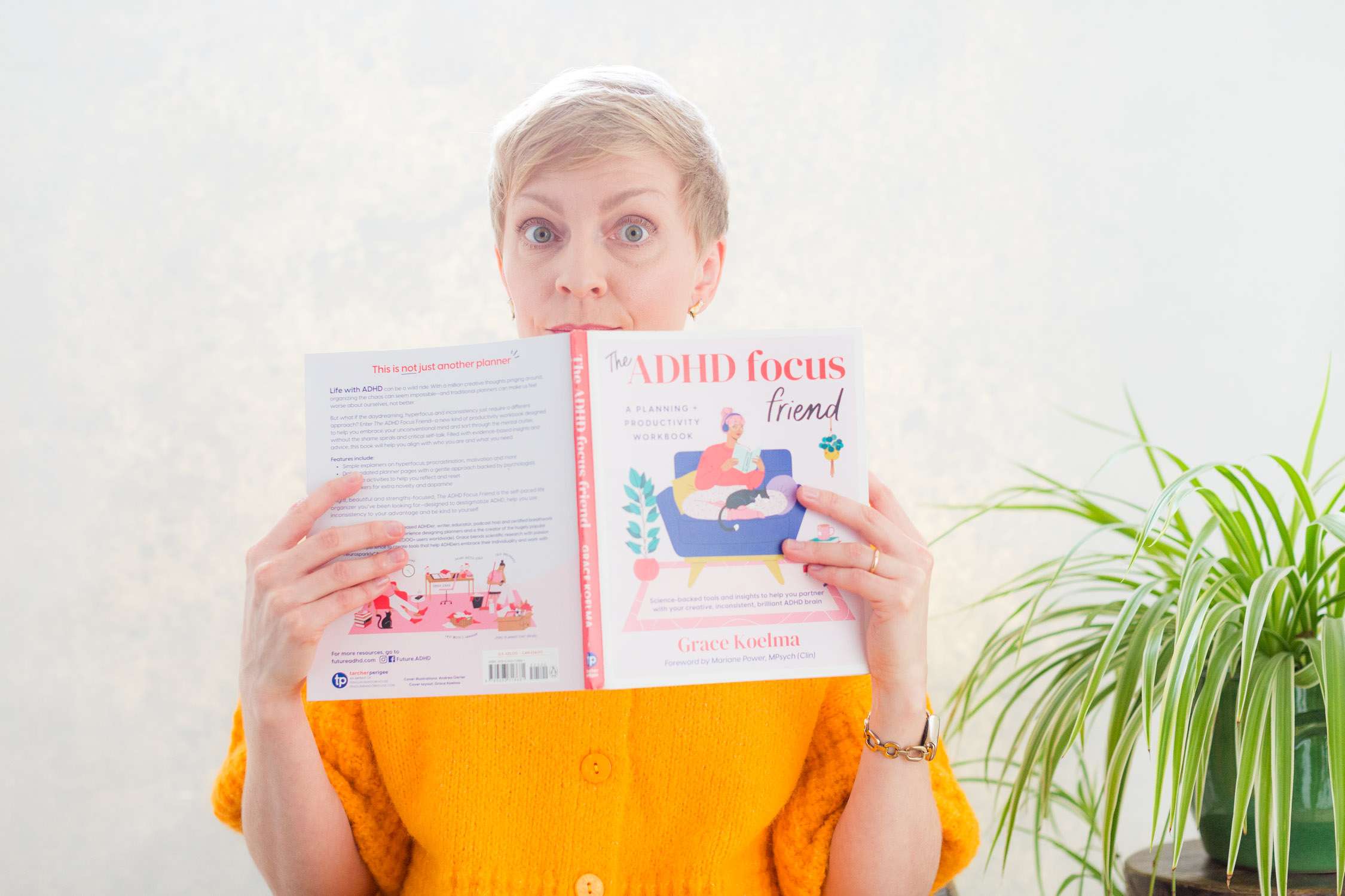 ADHD coach raises eyebrows jokingly above handbook in studio photoshoot with plant in background
