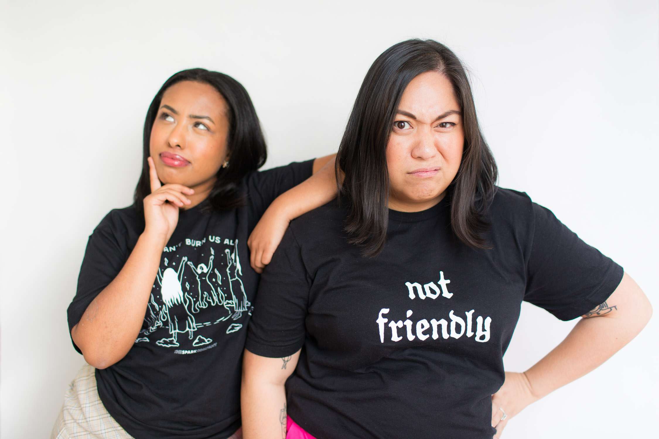 two comedians wearing black feminist t-shirts bearing slogan 'witches' - she looks comically pensive with an arm on shoulder of the comedian to the right, who has hands on hips and wears a 'not friendly' slogan t-shirt by the spark company