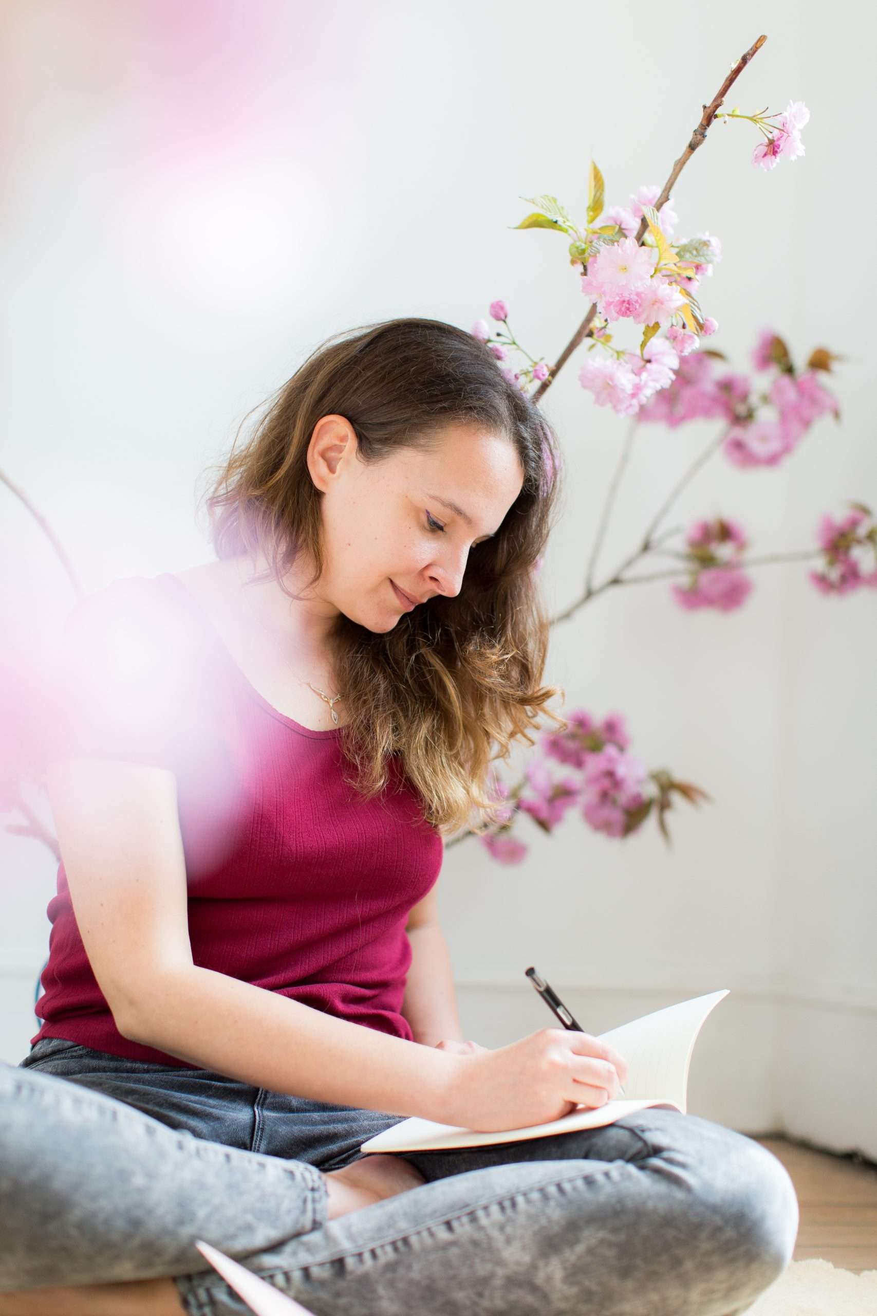 Jungian therapist sat cross legged in burgundy top and grey leggings sat amongst cherry blossom journalling while smiling