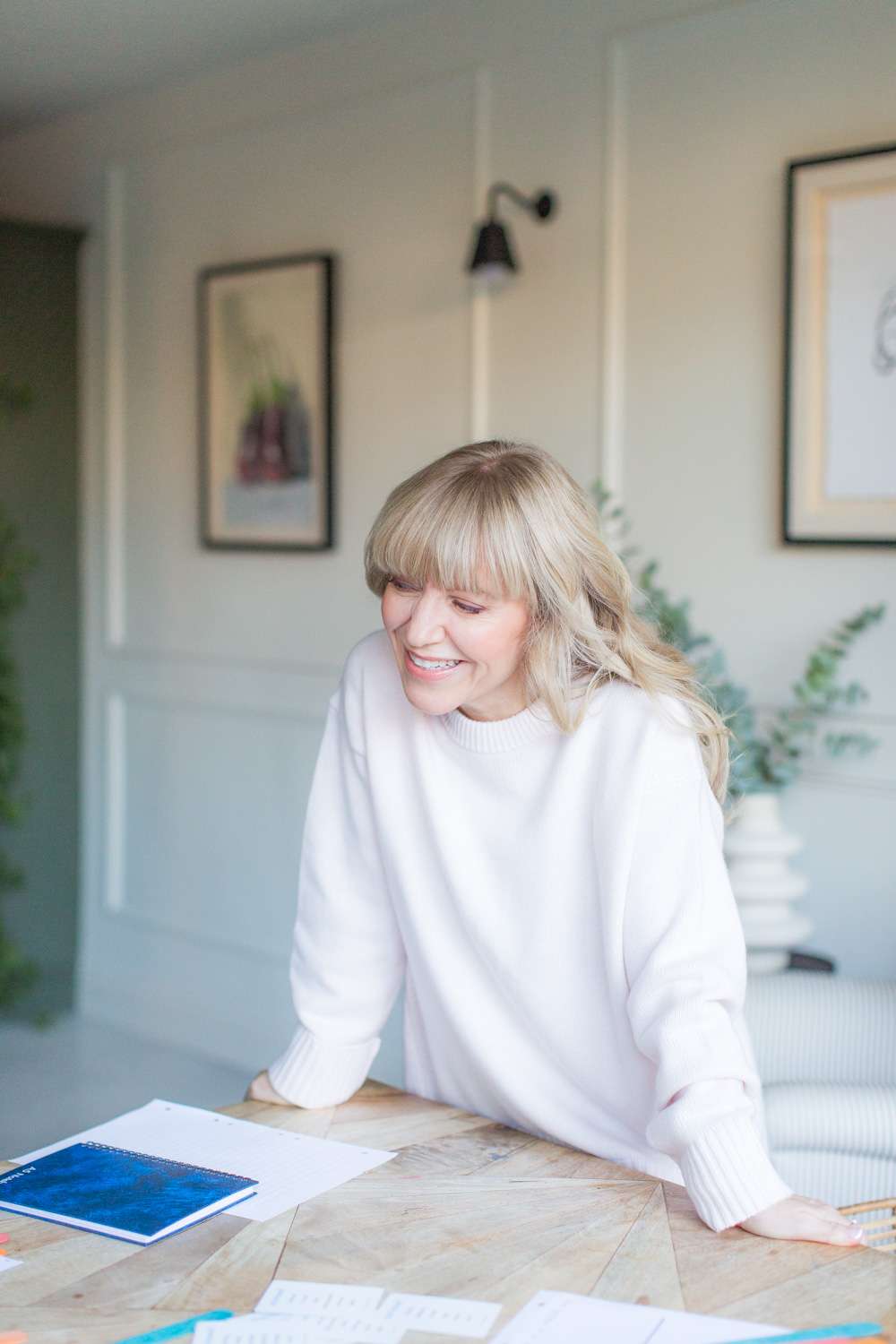 ash-blonde tutor wearing pale pink jumper leans, smiling, over table full of books, framed plant pictures in background