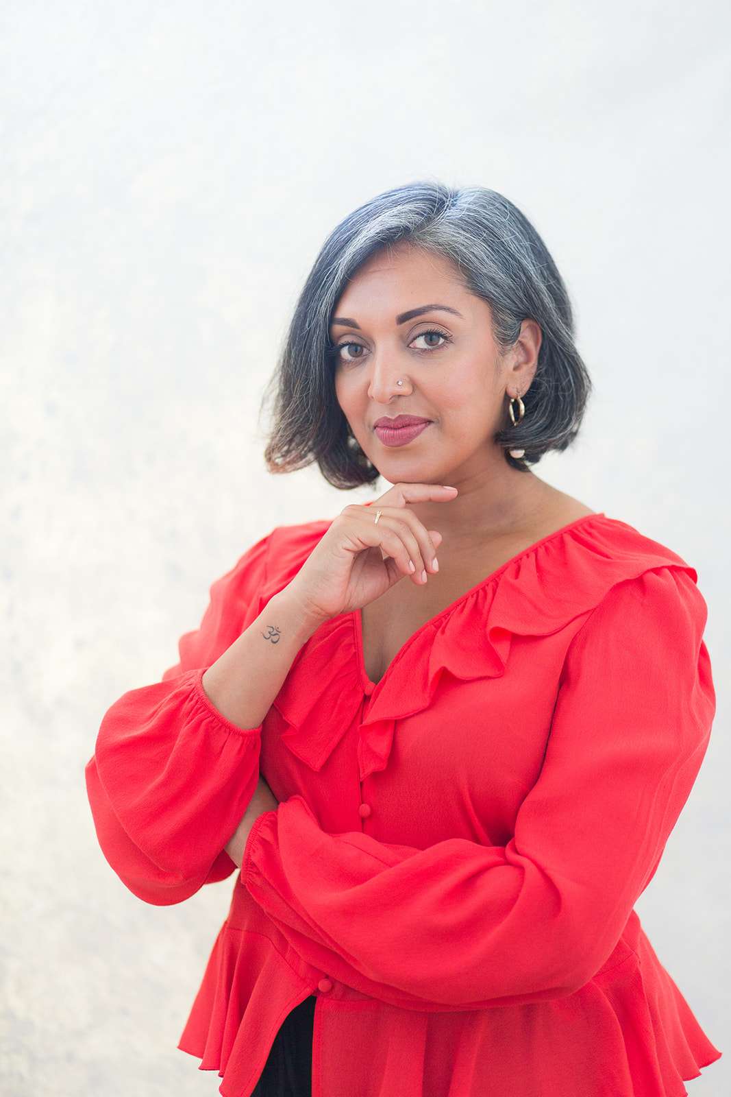 Headshot of female executive leadership coach dressed in coral long sleeved ruffled blouse and small gold hoop earrings, head raised looking directly into camera with her hand under her chin, left arm folded across body