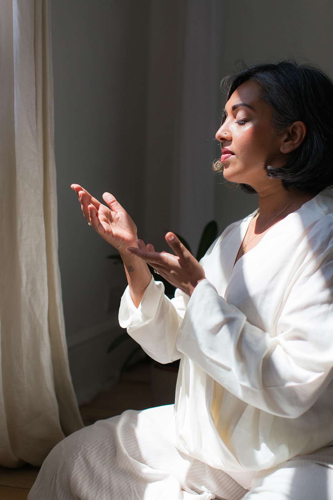 brand shoot photo of female reiki coach dressed all in white sat in sunlight strip in between shadows with hands raised to the light