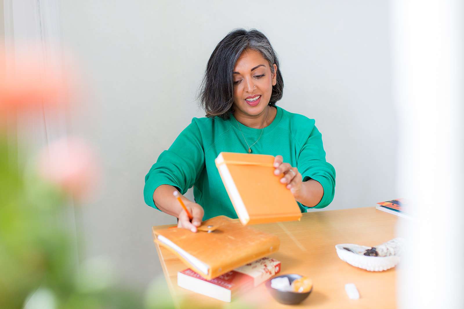 Peering from behind doorway and houseplant at a behind the scenes photo of a leadership coach dressed in a teal top picking up a journal from a stack on the table in front of her